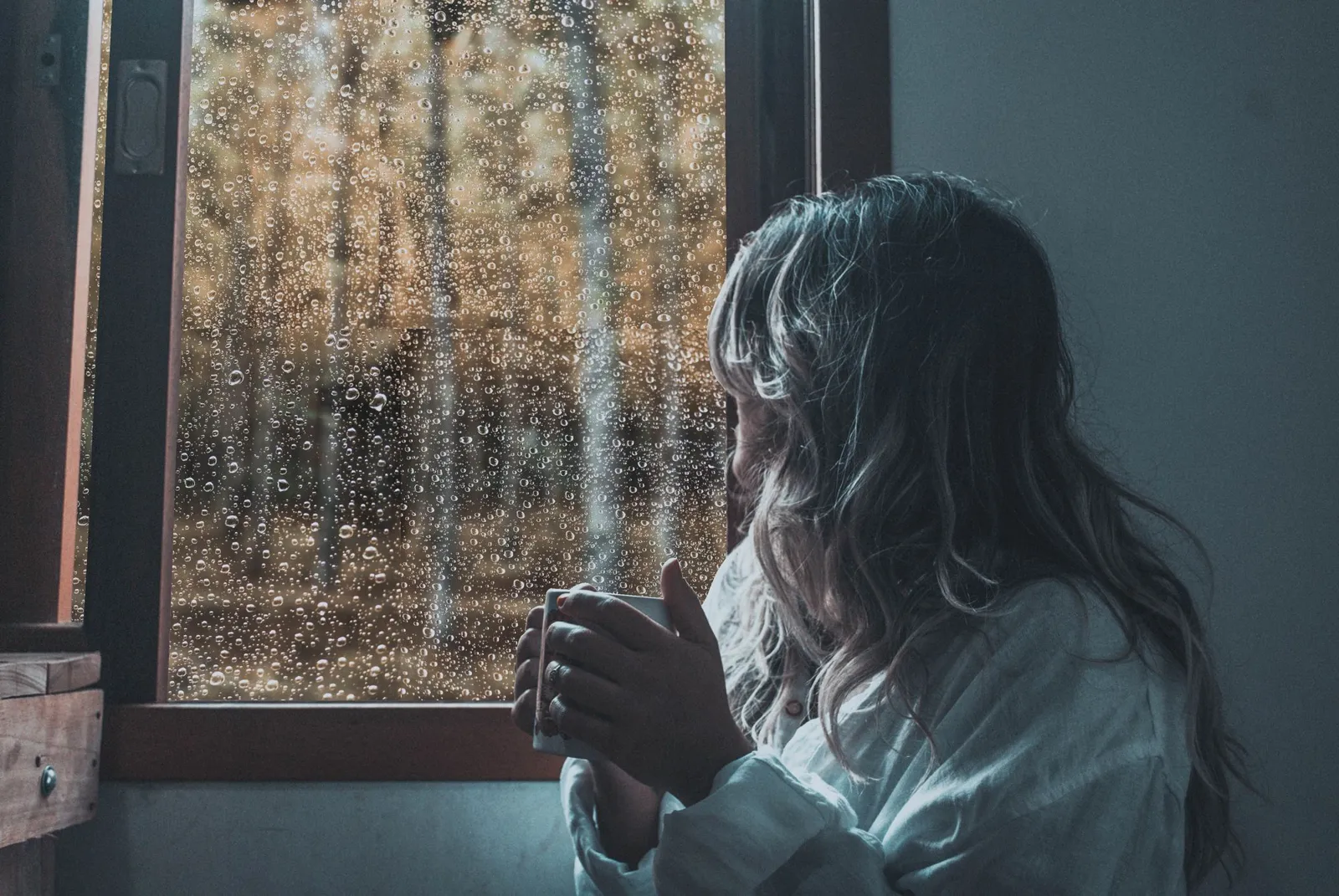 A woman holding a mug looks out of a rain-covered window